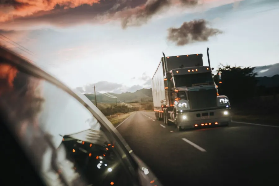 Semi-truck with its headlights on, on a highway at dusk